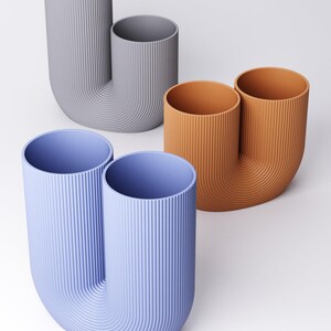 K&ouml;nnte beinhalten: Drei U-f&ouml;rmige Vasen mit vertikaler Rippenstruktur. Eine Vase ist hellblau, eine terrakottafarben und eine hellgrau. Jede Vase verf&uuml;gt &uuml;ber zwei zylindrische &Ouml;ffnungen, die durch einen geschwungenen Boden verbunden sind und eine Doppelrohrform bilden.