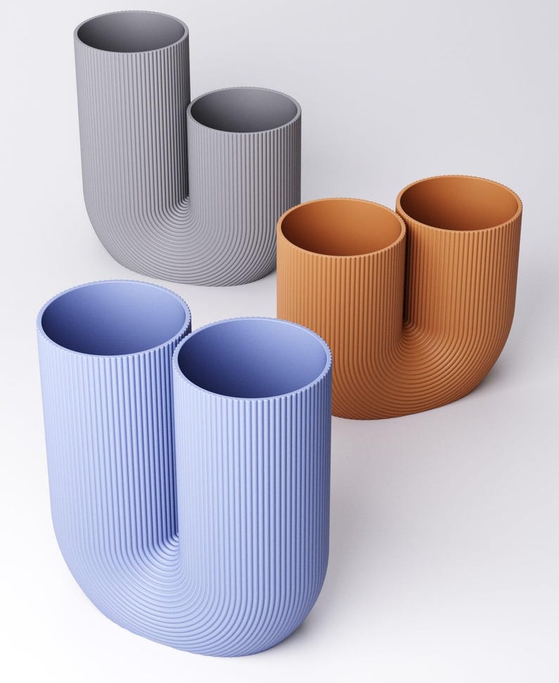 K&ouml;nnte beinhalten: Drei U-f&ouml;rmige Vasen mit vertikaler Rippenstruktur. Eine Vase ist hellblau, eine terrakottafarben und eine hellgrau. Jede Vase verf&uuml;gt &uuml;ber zwei zylindrische &Ouml;ffnungen, die durch einen geschwungenen Boden verbunden sind und eine Doppelrohrform bilden.