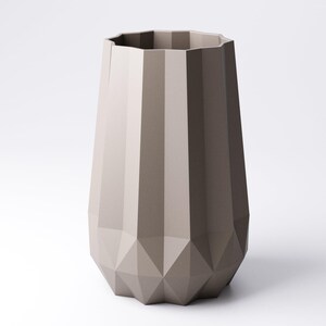K&ouml;nnte beinhalten: Eine taupefarbene Vase mit geometrischem Design. Die Vase hat eine facettierte, polygonale Form mit vertikalen Rippen und einem rautenf&ouml;rmigen Boden. Die Oberseite der Vase hat eine vielseitige &Ouml;ffnung. Die Vase steht vor einem wei&szlig;en Hintergrund.