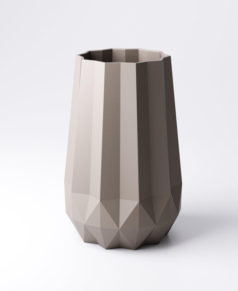 K&ouml;nnte beinhalten: Eine taupefarbene Vase mit geometrischem Design. Die Vase hat eine facettierte, polygonale Form mit vertikalen Rippen und einem rautenf&ouml;rmigen Boden. Die Oberseite der Vase hat eine vielseitige &Ouml;ffnung. Die Vase steht vor einem wei&szlig;en Hintergrund.