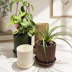Puede incluir: Cuatro macetas decorativas en tonos verdes, marrones y crema, cada una con un dise&ntilde;o acanalado. Las macetas est&aacute;n llenas de varias plantas verdes. Cada maceta se asienta sobre un platillo a juego. Una impresi&oacute;n bot&aacute;nica enmarcada est&aacute; en el fondo.