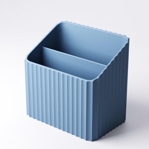 May include: A light blue desk organizer with a ribbed exterior and two interior compartments. The organizer is made of a solid material and has a rectangular shape with a slanted top. The design is simple and modern.