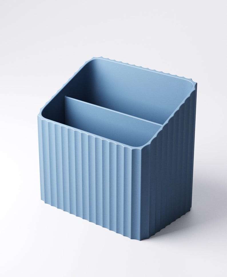 May include: A light blue desk organizer with a ribbed exterior and two interior compartments. The organizer is made of a solid material and has a rectangular shape with a slanted top. The design is simple and modern.