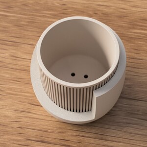 May include: A light beige, modern planter with a cylindrical interior and a decorative, vertical-slat design on the exterior. The planter has two drainage holes and sits on a wooden surface.