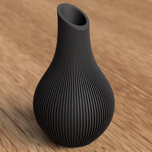May include: A black, textured vase with a unique, ribbed design. The vase has a rounded base and a slender, angled opening at the top. The vase is made of a dark material and sits on a wooden surface.