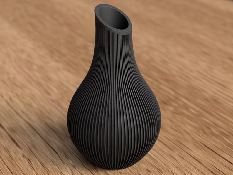 May include: A black, textured vase with a unique, ribbed design. The vase has a rounded base and a slender, angled opening at the top. The vase is made of a dark material and sits on a wooden surface.