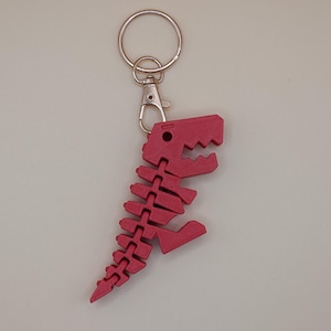 May include: A red dinosaur skeleton keychain. The keychain features a detailed, articulated design of a Tyrannosaurus Rex skeleton. The keychain is attached to a silver-colored split ring and a lobster clasp.
