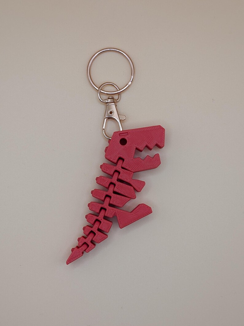 May include: A red dinosaur skeleton keychain. The keychain features a detailed, articulated design of a Tyrannosaurus Rex skeleton. The keychain is attached to a silver-colored split ring and a lobster clasp.