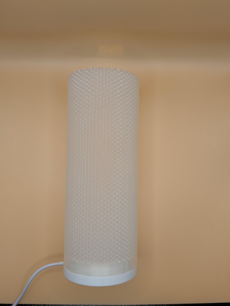 K&ouml;nnte beinhalten: Eine zylindrische Lampe mit einem strukturierten, gepunkteten Muster auf dem Lampenschirm. Die Lampe ist wei&szlig; und steht auf einem wei&szlig;en Sockel. Ein wei&szlig;es Kabel erstreckt sich von der Basis. Der Hintergrund ist einfarbig beige.