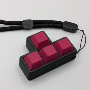 May include: A black, four-key mechanical keyboard with magenta keycaps. The keycaps are arranged in a Tetris-like shape. A black lanyard is attached to the keyboard. The background is white.