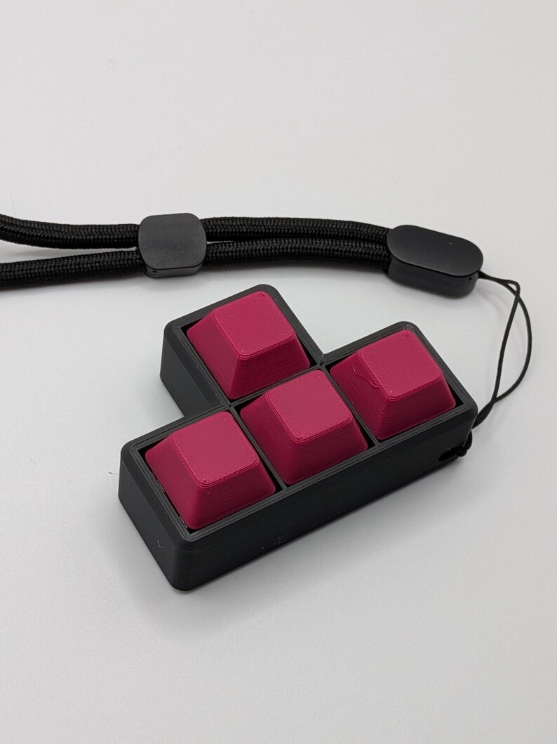 May include: A black, four-key mechanical keyboard with magenta keycaps. The keycaps are arranged in a Tetris-like shape. A black lanyard is attached to the keyboard. The background is white.