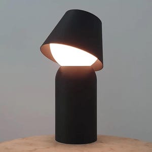 May include: A modern black table lamp with a unique design. The lamp features a cylindrical base and a tilted lampshade, revealing a bright, spherical light source. The interior of the shade has a gold finish.