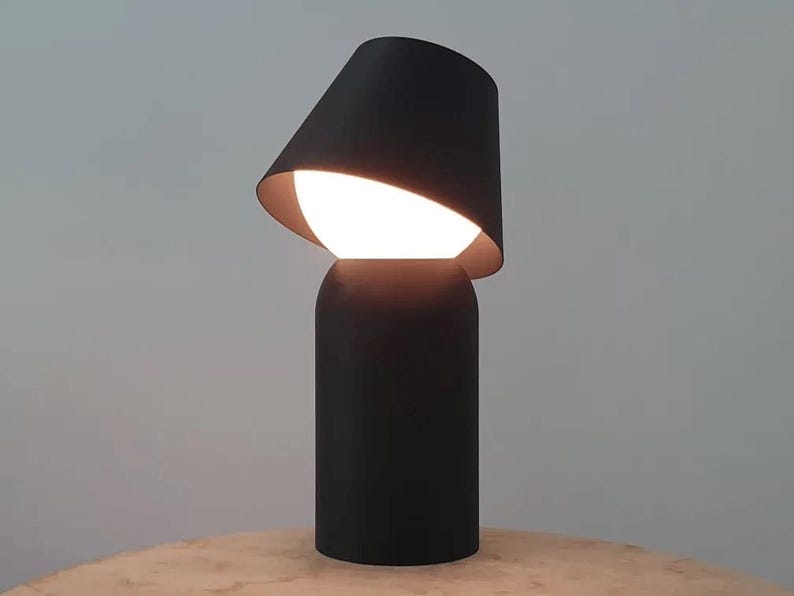 May include: A modern black table lamp with a unique design. The lamp features a cylindrical base and a tilted lampshade, revealing a bright, spherical light source. The interior of the shade has a gold finish.