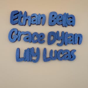 May include: Six blue glitter names: Ethan, Bella, Grace, Dylan, Lilly, and Lucas. The names are in a rounded font, suitable for nursery decor or personalised gifts. Each name has a unique size and shape.