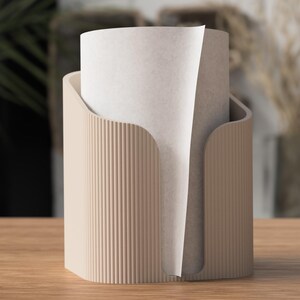 Op de afbeelding: Een beige papieren handdoekhouder met een verticaal geribbeld ontwerp. De houder heeft een gebogen opening om papieren handdoeken af te geven. Een rol papieren handdoeken bevindt zich in de houder.