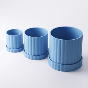 May include: Three blue cylindrical planters of varying sizes, each with a matching base. The planters have vertical ribbed detailing, creating a textured surface. The set includes a small, medium, and large planter, suitable for indoor or outdoor use.