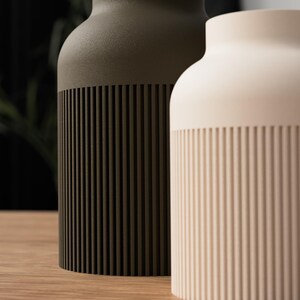 May include: Two ceramic vases, one olive green and the other cream-colored, sit side-by-side. The lower portion of each vase features vertical, grooved detailing. The vases have a rounded shape and are displayed on a wooden surface.