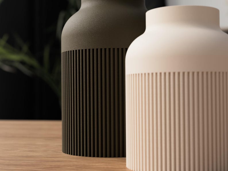 May include: Two ceramic vases, one olive green and the other cream-colored, sit side-by-side. The lower portion of each vase features vertical, grooved detailing. The vases have a rounded shape and are displayed on a wooden surface.