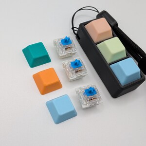 May include: A collection of colorful keyboard keycaps in teal, orange, and light blue, along with clear mechanical keyboard switches with blue accents. A black holder contains three keycaps in peach, light green, and light blue. A black wrist strap is attached.