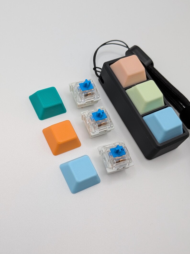 May include: A collection of colorful keyboard keycaps in teal, orange, and light blue, along with clear mechanical keyboard switches with blue accents. A black holder contains three keycaps in peach, light green, and light blue. A black wrist strap is attached.