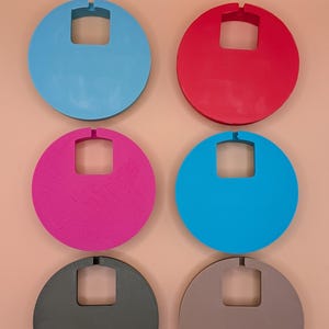 K&ouml;nnte beinhalten: Sechs farbige, kreisf&ouml;rmige Objekte mit einem quadratischen Ausschnitt und einer kleinen Kerbe oben. Die Farben sind Blau, Rot, Pink, T&uuml;rkis, Grau und Braun. Die Objekte sind in zwei Reihen zu je drei St&uuml;ck auf einem pfirsichfarbenen Hintergrund angeordnet.