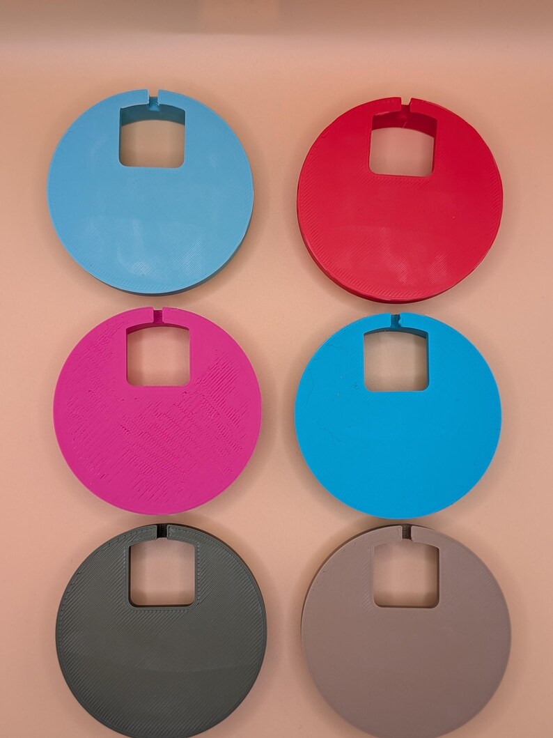 K&ouml;nnte beinhalten: Sechs farbige, kreisf&ouml;rmige Objekte mit einem quadratischen Ausschnitt und einer kleinen Kerbe oben. Die Farben sind Blau, Rot, Pink, T&uuml;rkis, Grau und Braun. Die Objekte sind in zwei Reihen zu je drei St&uuml;ck auf einem pfirsichfarbenen Hintergrund angeordnet.