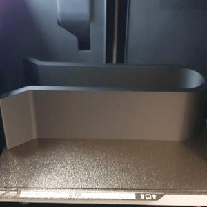 K&ouml;nnte beinhalten: Bauplatte eines 3D-Druckers mit einem grauen, rechteckigen Objekt. Das Objekt hat eine gebogene Oberseite und eine flache Basis, wahrscheinlich zur Aufnahme eines bestimmten Gegenstands w&auml;hrend des Druckvorgangs. Das Innere des Druckers ist sichtbar.