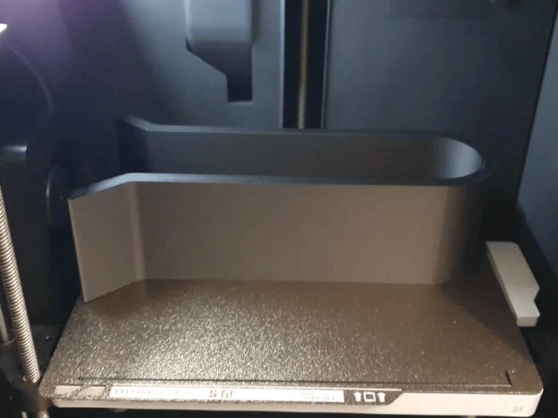 K&ouml;nnte beinhalten: Bauplatte eines 3D-Druckers mit einem grauen, rechteckigen Objekt. Das Objekt hat eine gebogene Oberseite und eine flache Basis, wahrscheinlich zur Aufnahme eines bestimmten Gegenstands w&auml;hrend des Druckvorgangs. Das Innere des Druckers ist sichtbar.