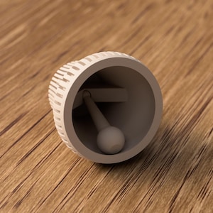 May include: A light gray bell with a spherical clapper, lying on a wooden surface. The bell's exterior has a textured, slotted design. The clapper is attached to the bell's interior.