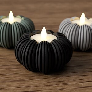 May include: Three decorative pumpkin-shaped candle holders in shades of black, gray, and green. Each holder has a ribbed design and holds a small, glowing LED candle. The holders are arranged on a wooden surface.