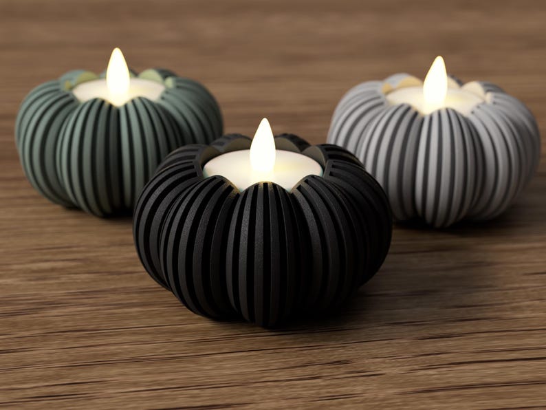 May include: Three decorative pumpkin-shaped candle holders in shades of black, gray, and green. Each holder has a ribbed design and holds a small, glowing LED candle. The holders are arranged on a wooden surface.