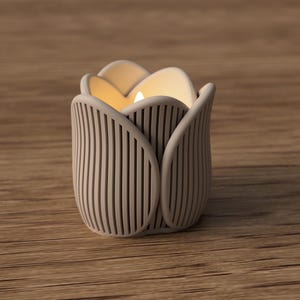 May include: A beige tulip-shaped candle holder with vertical lines and a lit candle inside. The candle holder is made of a light-colored material and sits on a wooden surface. The candle flame glows warmly.