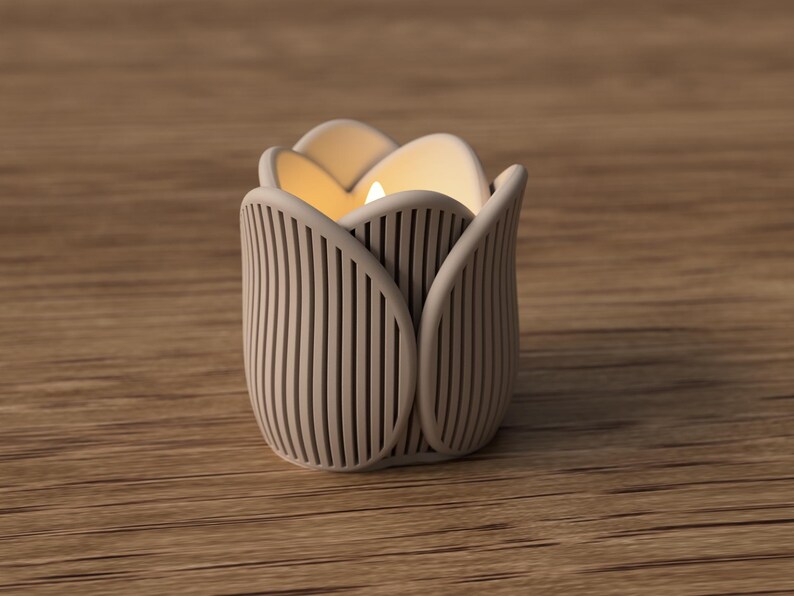 May include: A beige tulip-shaped candle holder with vertical lines and a lit candle inside. The candle holder is made of a light-colored material and sits on a wooden surface. The candle flame glows warmly.