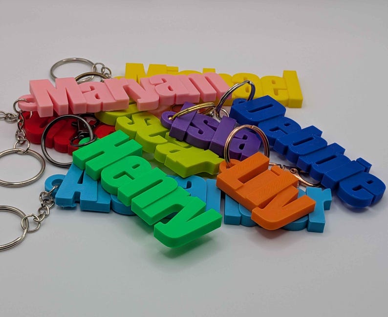 May include: A collection of colorful 3D-printed name keychains in various colors, including pink, yellow, green, blue, and orange. Each keychain features a different name and includes a metal keyring.