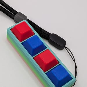 May include: A rectangular, handheld device with four square buttons in red and blue. The device has a light blue frame with a gradient of colors along the edges. A black lanyard is attached to the top.