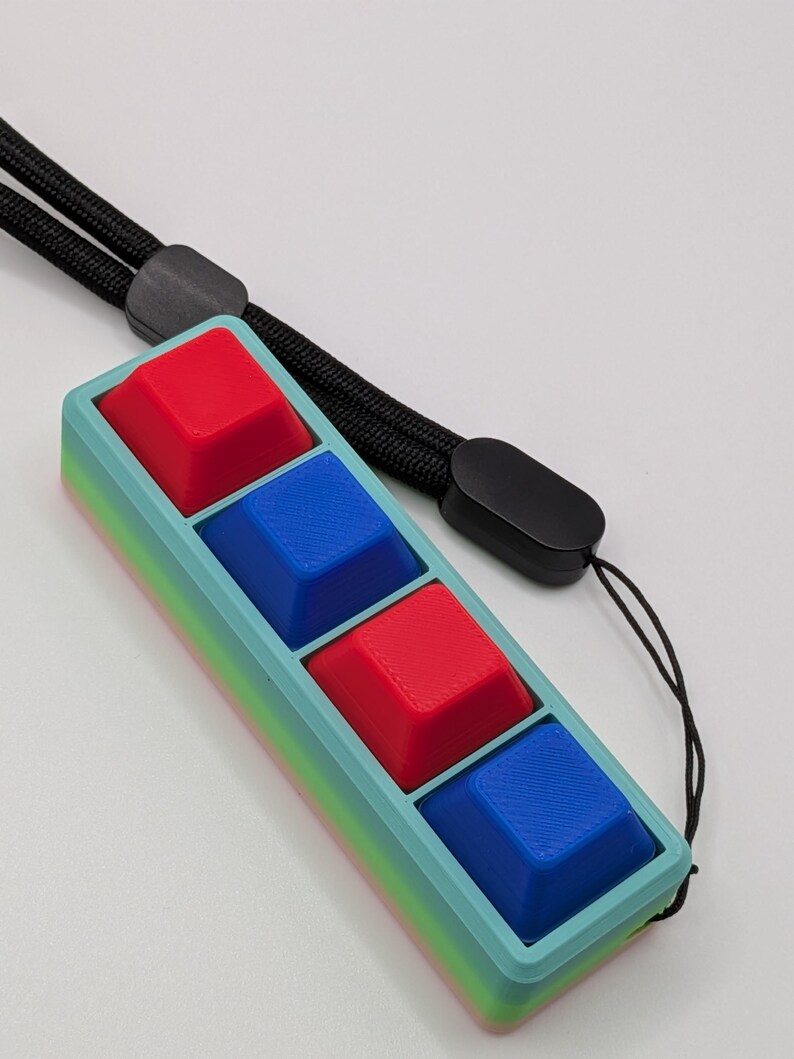 May include: A rectangular, handheld device with four square buttons in red and blue. The device has a light blue frame with a gradient of colors along the edges. A black lanyard is attached to the top.