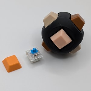 May include: A black sphere with beige, brown, and light pink square keycaps. An orange keycap and a clear mechanical keyboard switch with a blue stem are next to the sphere. The sphere is a unique design for a keyboard.