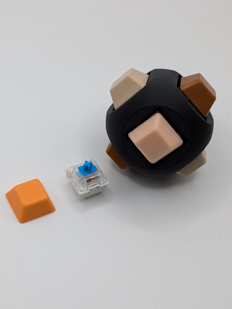 May include: A black sphere with beige, brown, and light pink square keycaps. An orange keycap and a clear mechanical keyboard switch with a blue stem are next to the sphere. The sphere is a unique design for a keyboard.