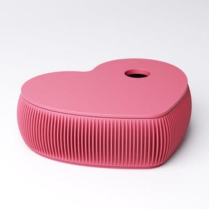 May include: A pink heart-shaped box with a lid. The box has a ribbed design around the base and a circular hole on the lid. The box is a solid, vibrant pink color and sits on a white background.
