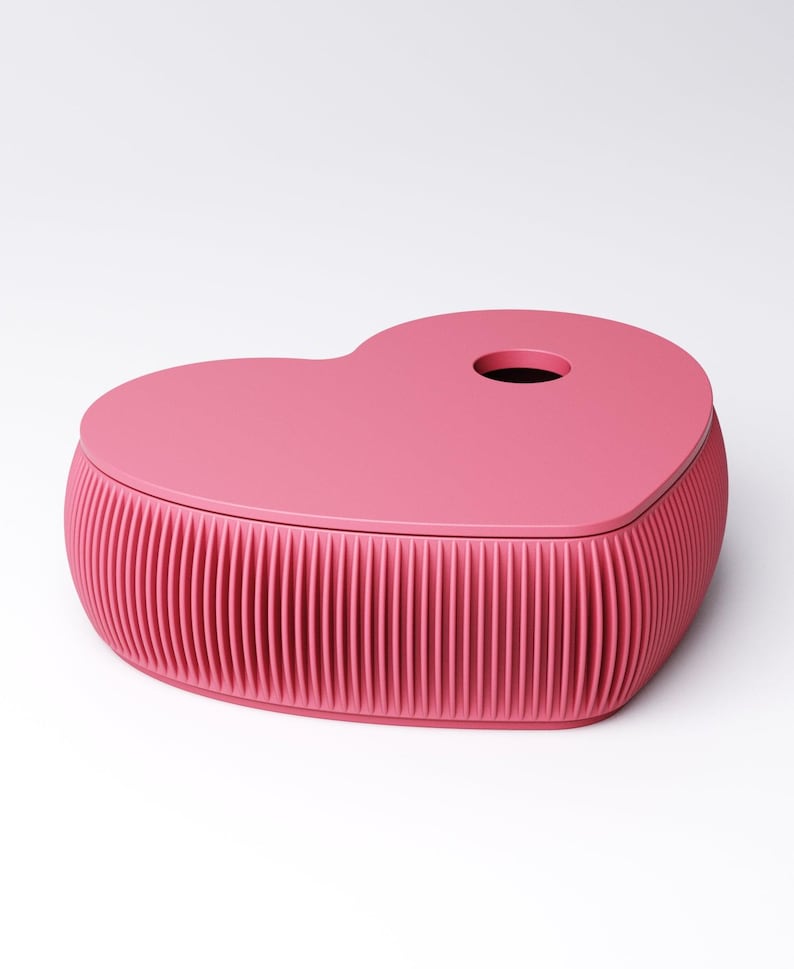 May include: A pink heart-shaped box with a lid. The box has a ribbed design around the base and a circular hole on the lid. The box is a solid, vibrant pink color and sits on a white background.