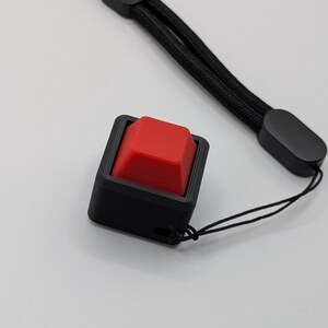 May include: A black square button with a red square button top, attached to a black lanyard. The button is in a black square frame. The lanyard is made of black woven material.