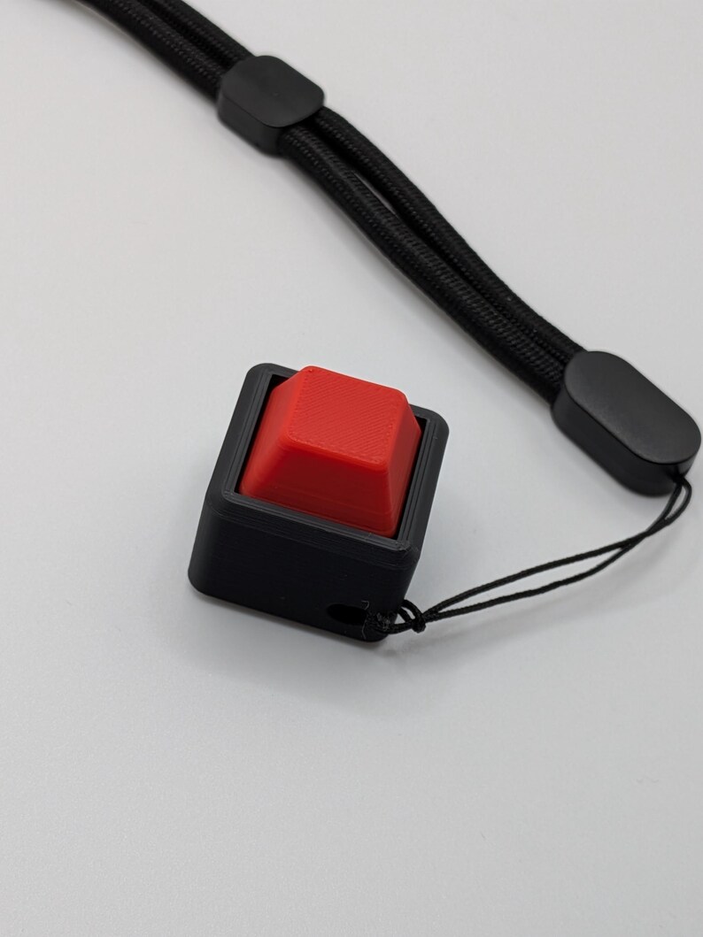 May include: A black square button with a red square button top, attached to a black lanyard. The button is in a black square frame. The lanyard is made of black woven material.