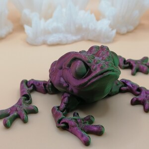 May include: A 3D-printed frog toy in shades of purple and green, with articulated limbs. The frog is positioned on a light beige surface, with white, crystalline formations in the background. The frog's eyes are large and detailed.