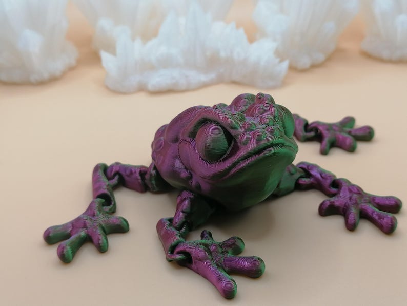 May include: A 3D-printed frog toy in shades of purple and green, with articulated limbs. The frog is positioned on a light beige surface, with white, crystalline formations in the background. The frog's eyes are large and detailed.