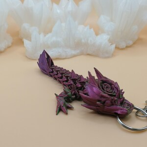 May include: A purple and green dragon keychain with a silver clasp and ring. The dragon has a detailed, textured design with a rose on its head. White crystal formations are in the background.