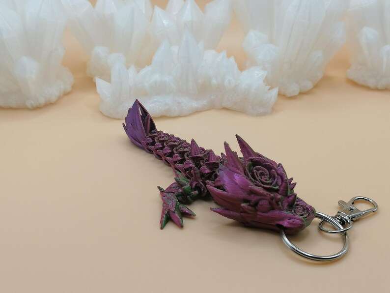 May include: A purple and green dragon keychain with a silver clasp and ring. The dragon has a detailed, textured design with a rose on its head. White crystal formations are in the background.