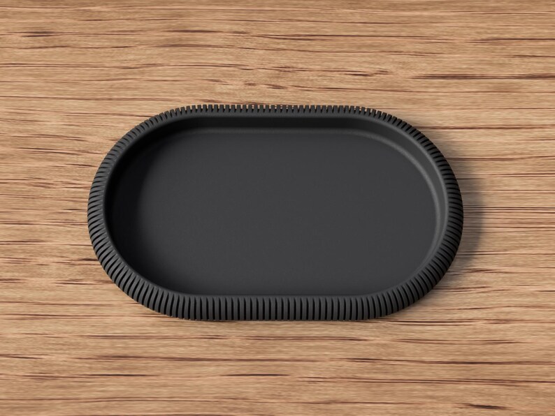Puede incluir: Una bandeja ovalada negra mate con un borde texturizado y acanalado. La bandeja est&aacute; vac&iacute;a y descansa sobre una superficie de madera. El dise&ntilde;o es simple y moderno, adecuado para servir o exhibir.