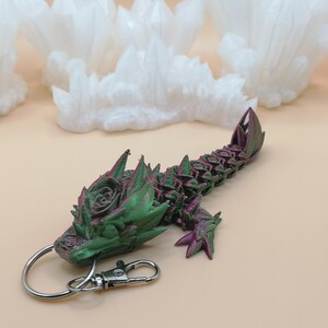May include: A 3D-printed dragon keychain in shades of green and purple. The dragon features a detailed design with a rose-like pattern on its head and back. It has a silver keyring and clasp for easy attachment.