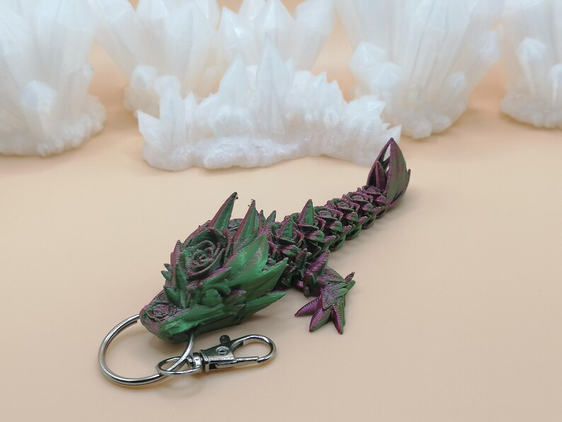 May include: A 3D-printed dragon keychain in shades of green and purple. The dragon features a detailed design with a rose-like pattern on its head and back. It has a silver keyring and clasp for easy attachment.