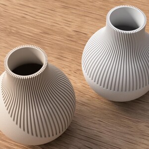 May include: Two off-white, modern vases with a ribbed design. The vases have a rounded base and a cylindrical opening. The ribbed pattern covers the body of the vases, creating a textured look. They sit on a light brown wooden surface.
